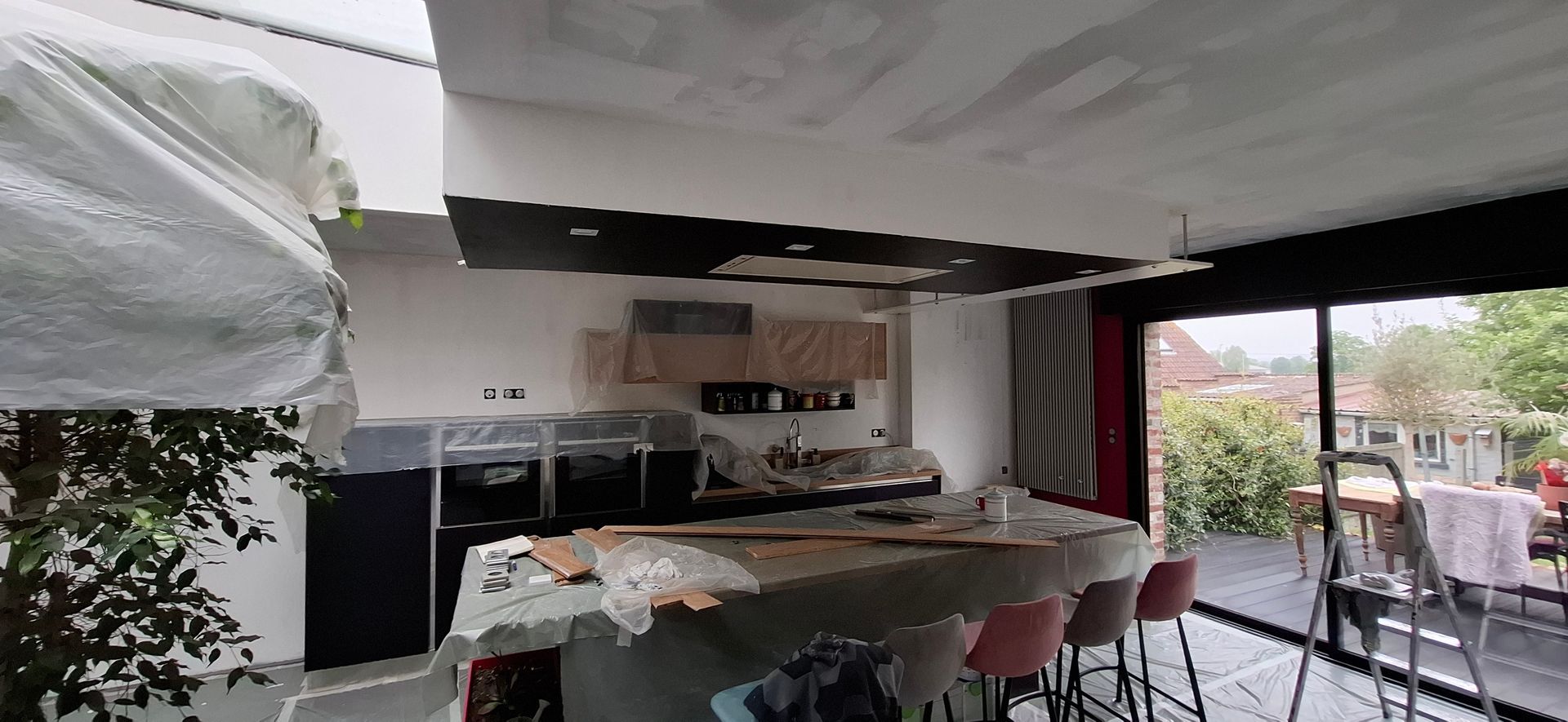 Une cuisine en construction avec un îlot central, des tabourets de bar et un grand plafond noir, visible à travers une vitre.