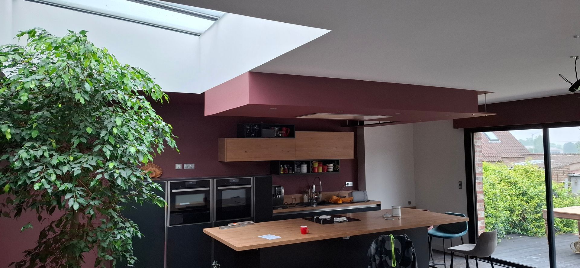 Intérieur de cuisine moderne avec des armoires foncées, un îlot en bois, une grande plante en pot et un puits de lumière sous un plafond à soffite rouge.