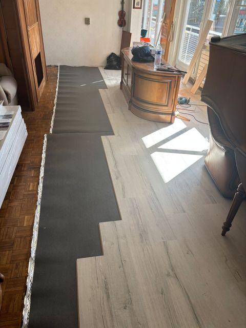 Un sol en rénovation montrant des bandes de sous-couche gris foncé recouvrant partiellement un parquet clair dans une pièce.