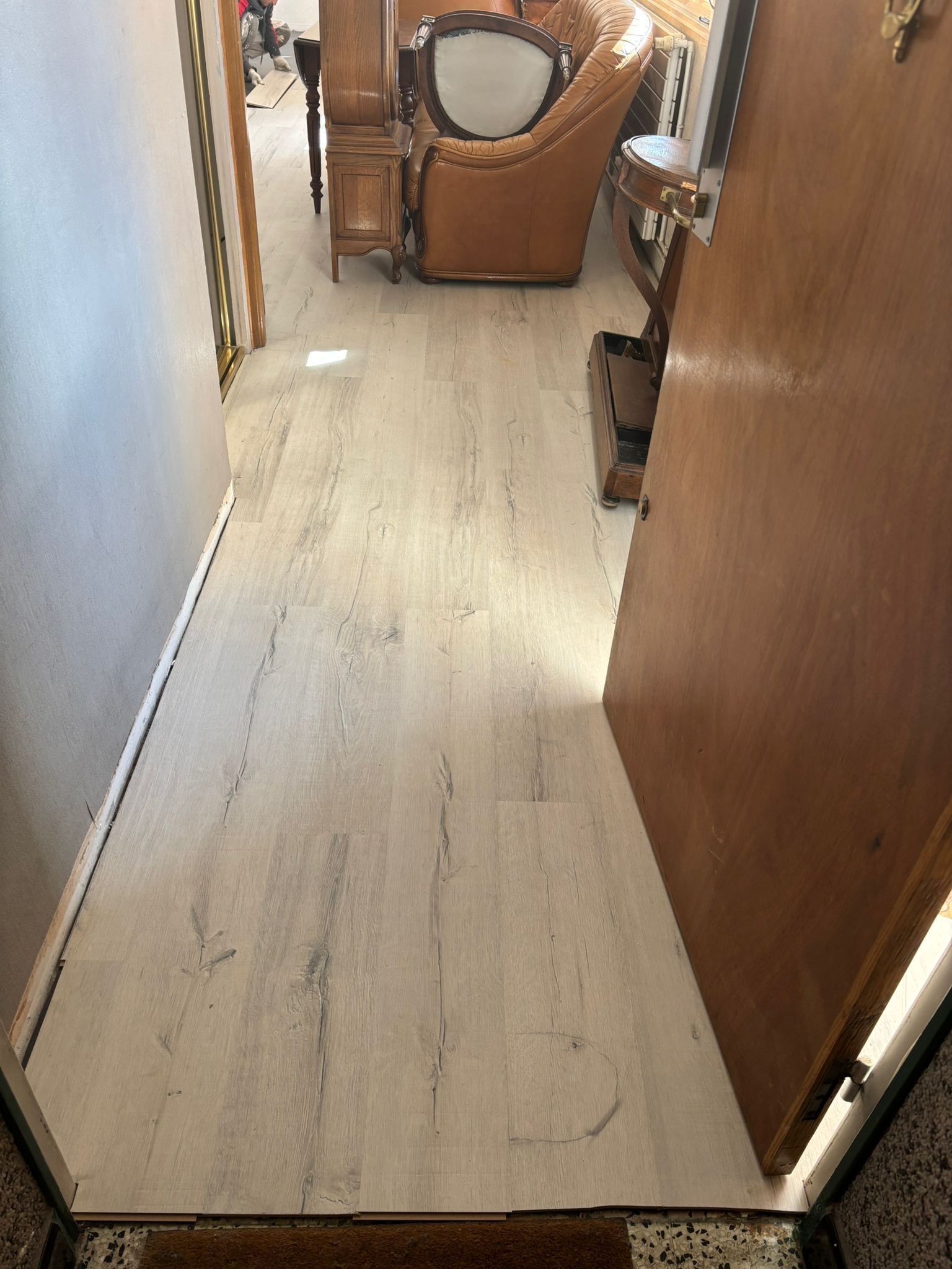 Un parquet clair orne un couloir étroit menant à une pièce où se trouve une chaise marron.