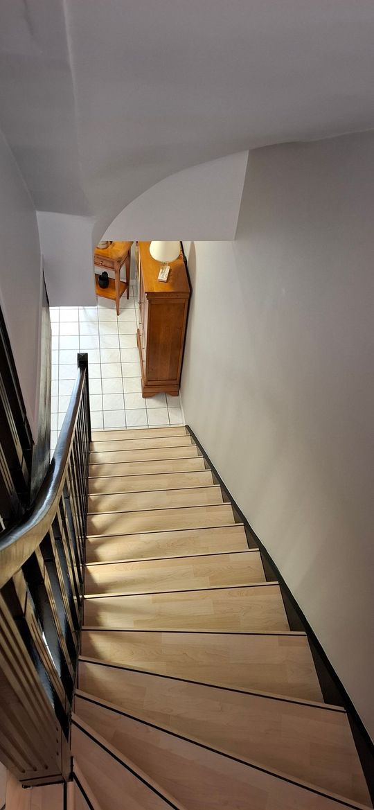 Vue plongeante d'un escalier en bois avec une rampe en métal noir descendant vers un couloir carrelé avec une armoire en bois.