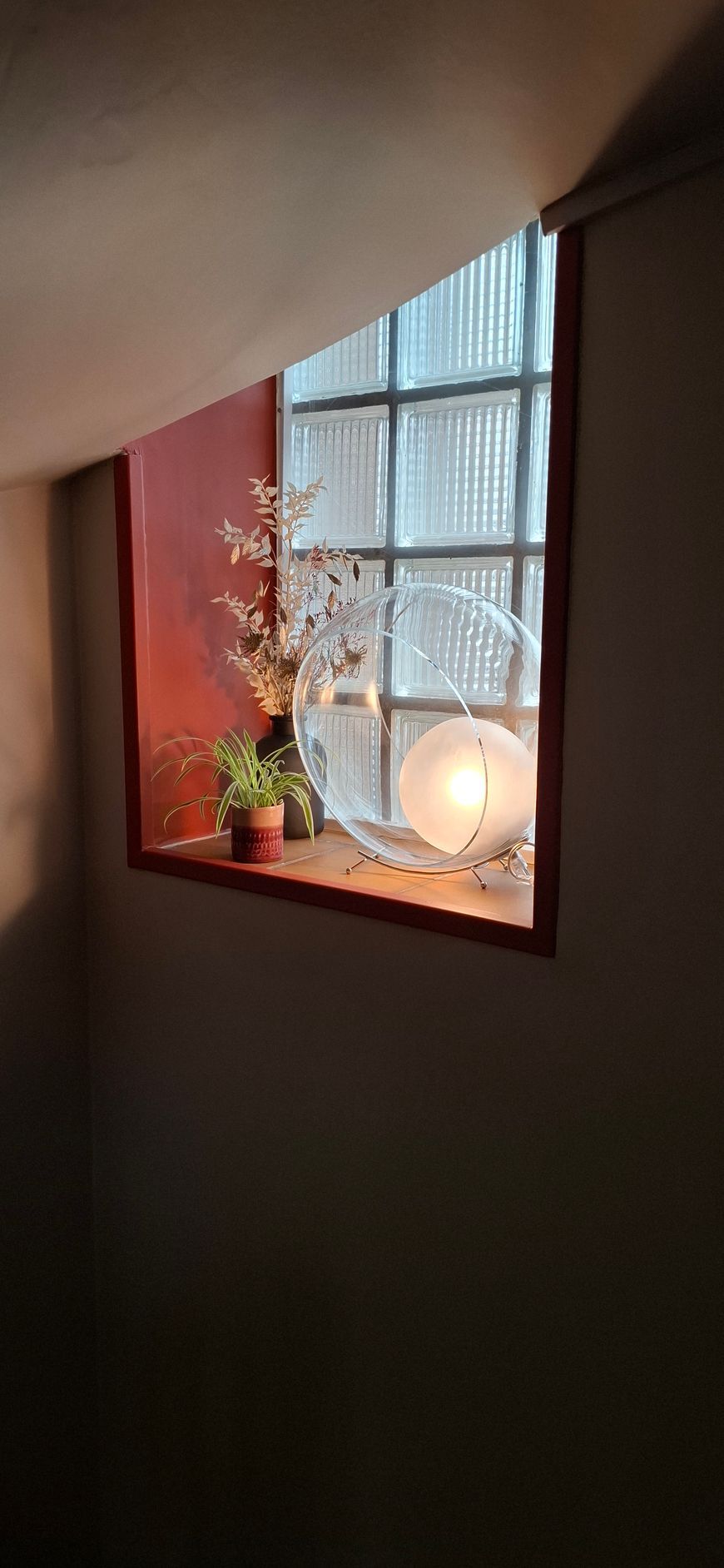Un rebord de fenêtre décoratif avec une plante en pot, un petit vase de fleurs et une lampe en forme de lune illuminée contre une vitre.