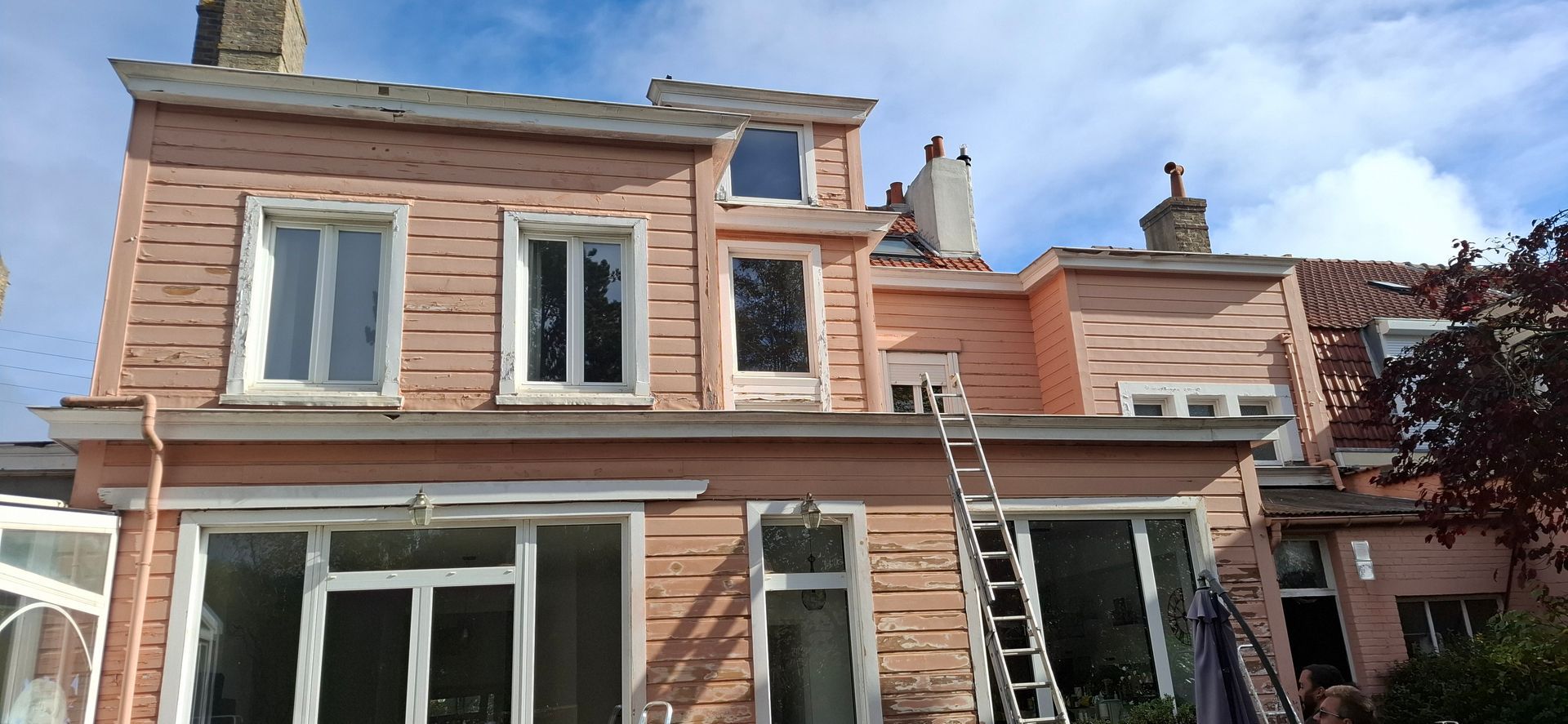 Une maison à deux étages avec un bardage en bois couleur saumon, de grandes fenêtres et une échelle métallique appuyée contre le mur extérieur.