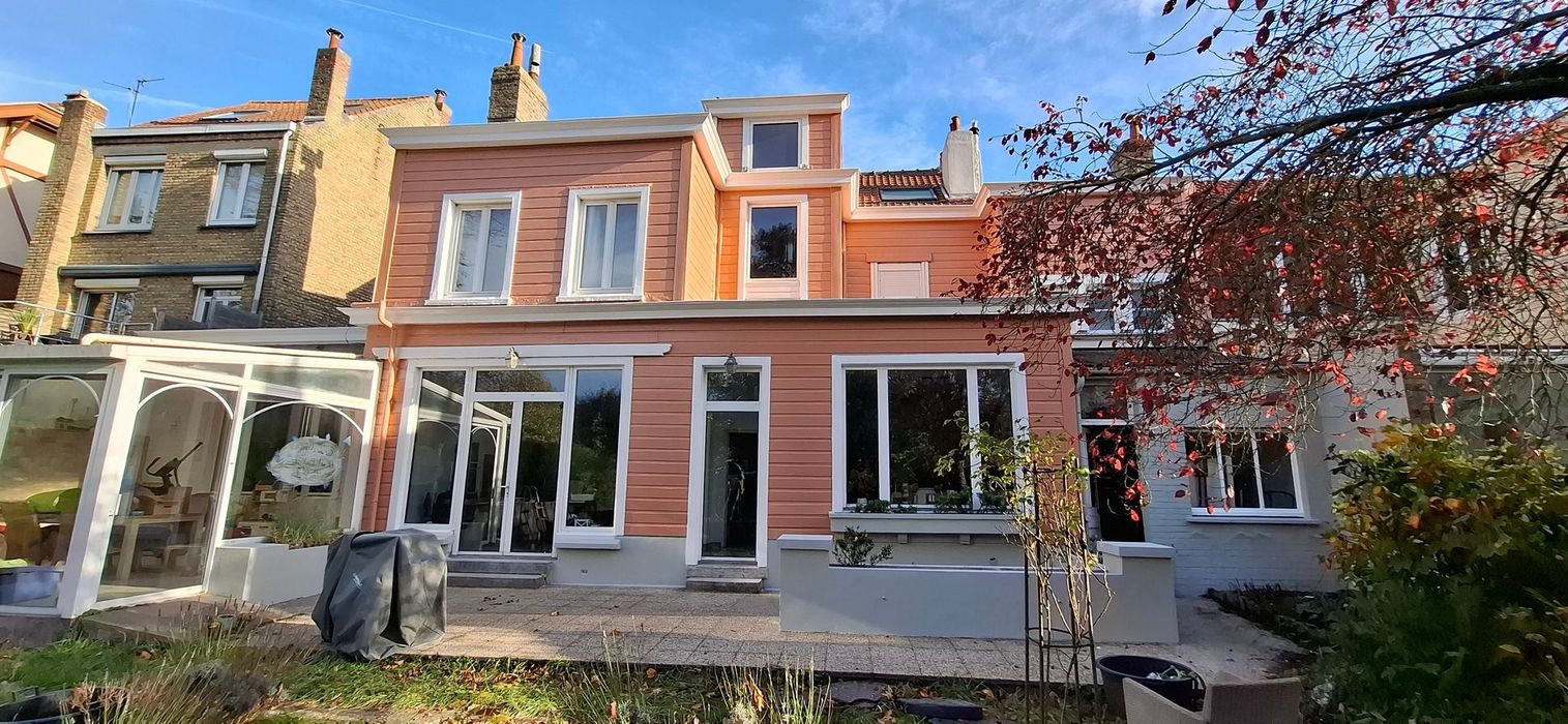 Une maison mitoyenne à plusieurs étages, couleur saumon, avec des encadrements de fenêtres blancs et un patio-jardin à l'arrière, sous un ciel d'un bleu éclatant.