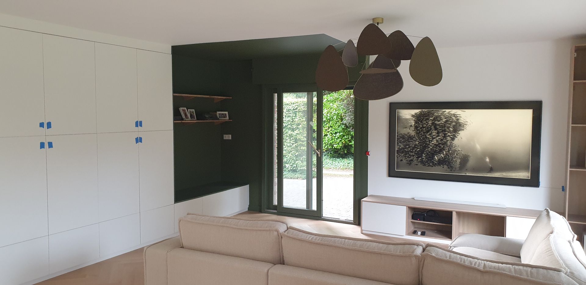 Salon avec mur d'accent vert olive, rangements intégrés blancs et grande télévision. Canapé d'angle beige.