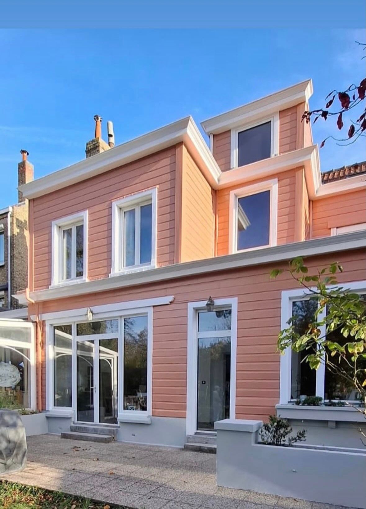Une maison de deux étages couleur pêche, avec des boiseries blanches, de grandes fenêtres et une terrasse en gravier sous un ciel bleu azur.