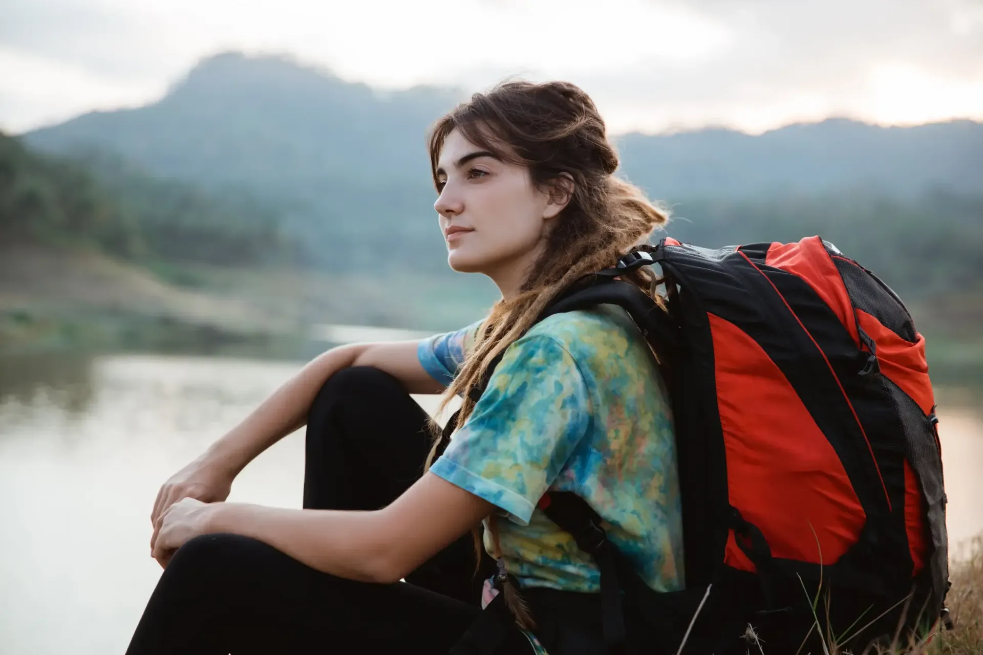 Una mujer con una mochila se sienta junto al agua, mirando una cadena montañosa.