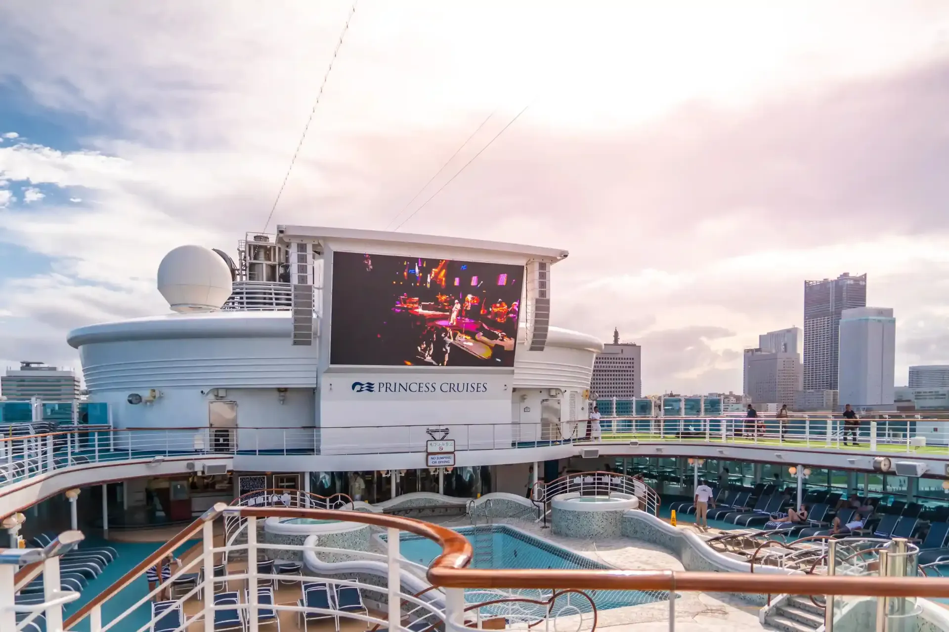 Cubierta de crucero con piscina, pantalla grande, paisaje urbano de fondo, cielo nublado.