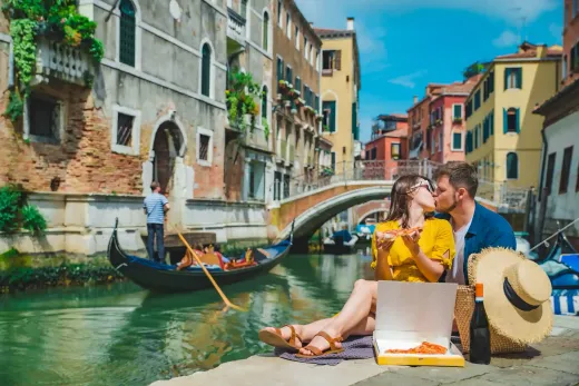Pareja besándose y comiendo pizza en Venecia, cerca de un canal con góndola; edificios y puente al fondo.