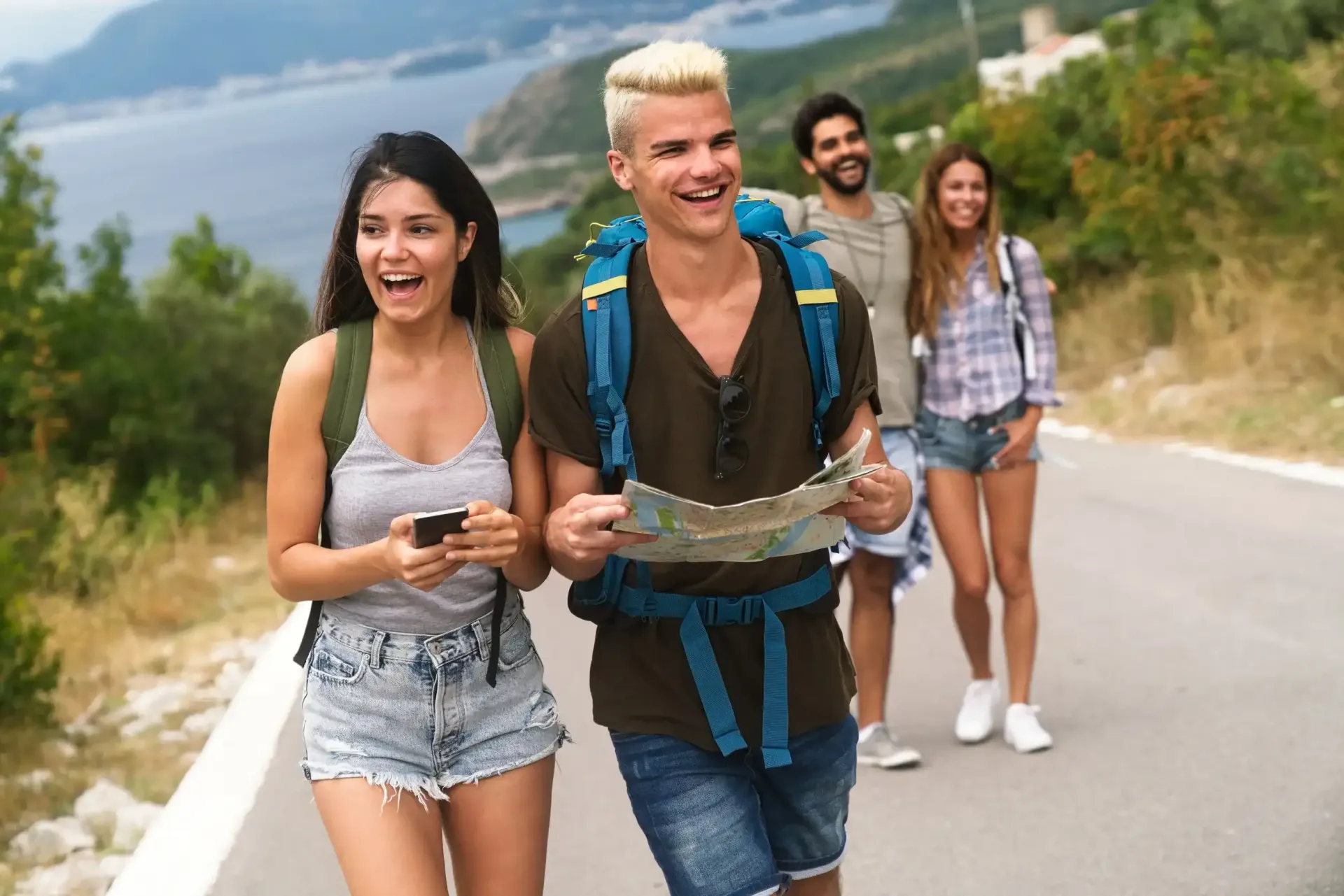 Grupo de amigos caminando por una carretera cerca de un cuerpo de agua, sonriendo, uno sosteniendo un mapa y otro un teléfono.