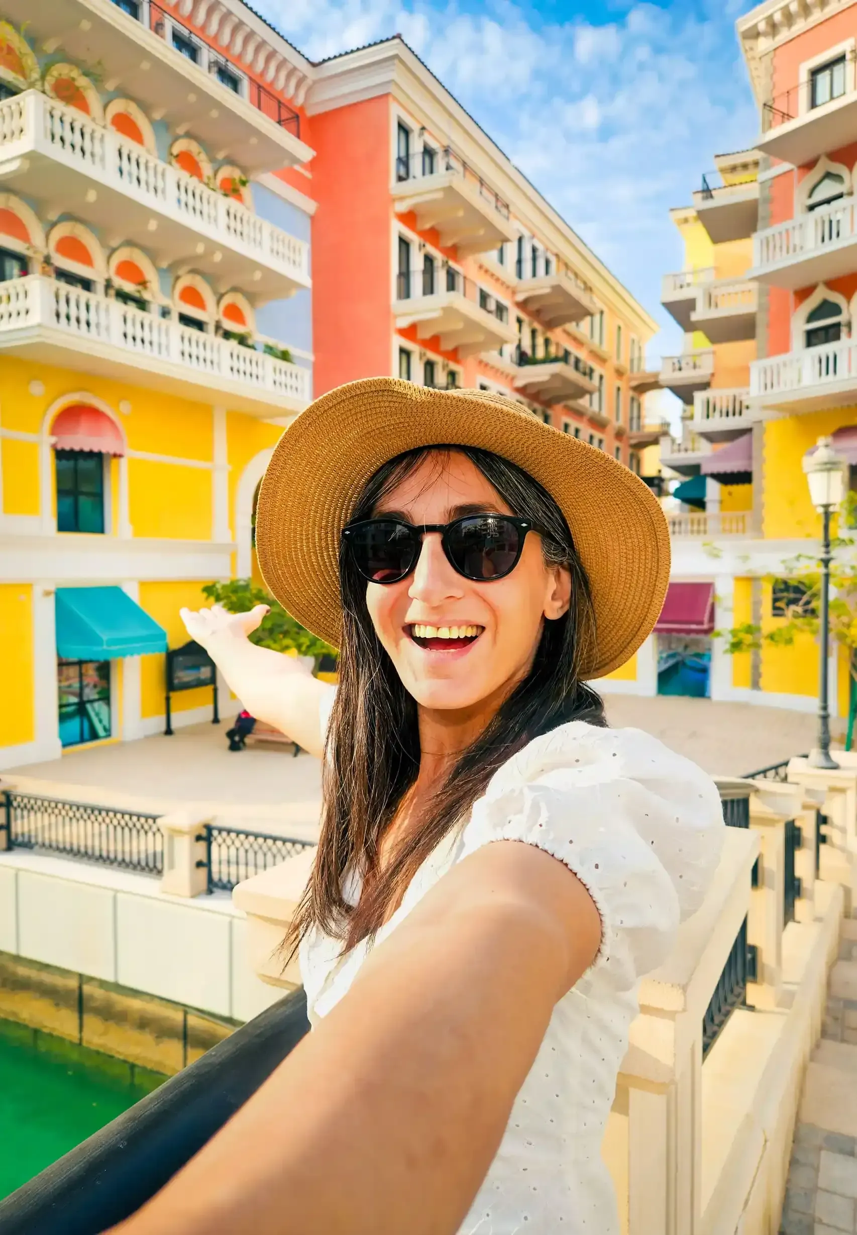 Una mujer con gafas de sol y sombrero sonríe y señala hacia edificios coloridos con arcos y balcones.