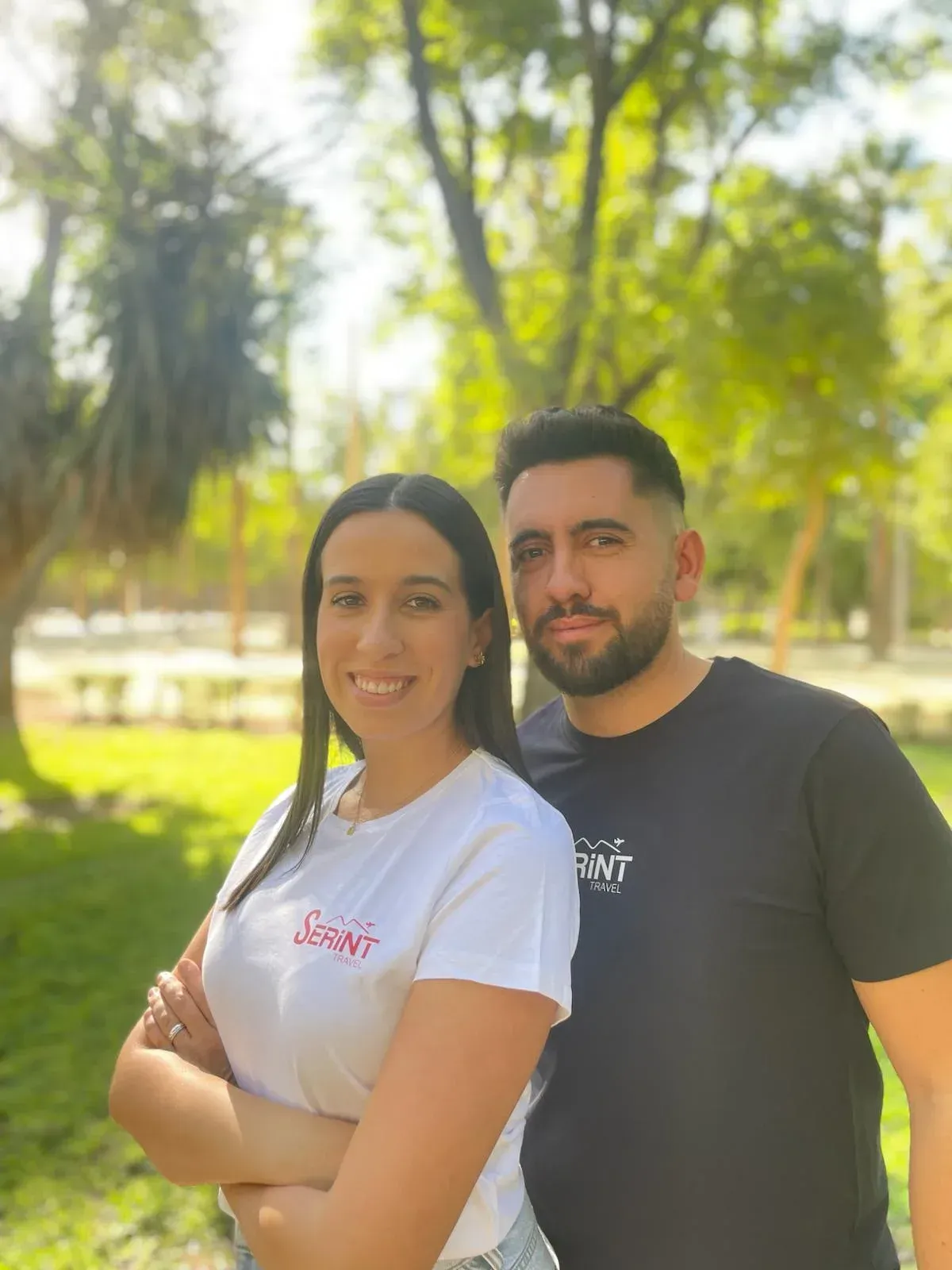 Dos personas sonriendo al aire libre; mujer con camisa blanca con los brazos cruzados, hombre con camisa negra.
