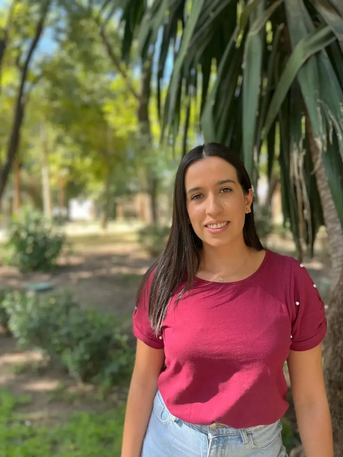 Una mujer de cabello oscuro sonríe, vistiendo una camisa burdeos y jeans, parada afuera cerca del follaje.