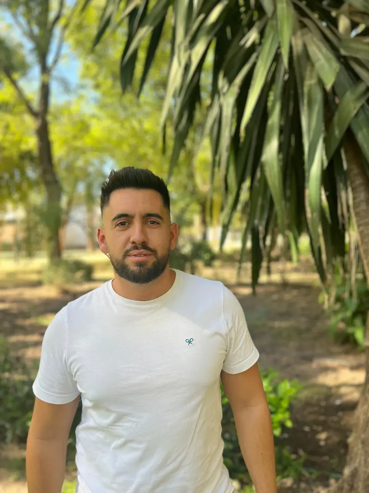 Hombre con camisa blanca sonriendo al aire libre, de pie cerca de la vegetación, mirando a la cámara.