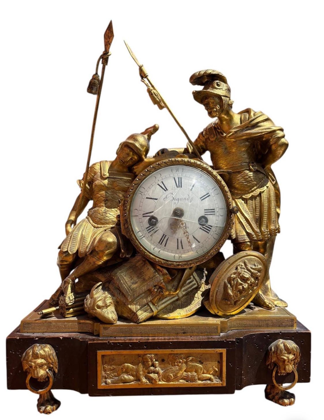 Horloge dorée ornée de figures de soldats romains brandissant des lances. Le cadran rond affiche des chiffres romains.