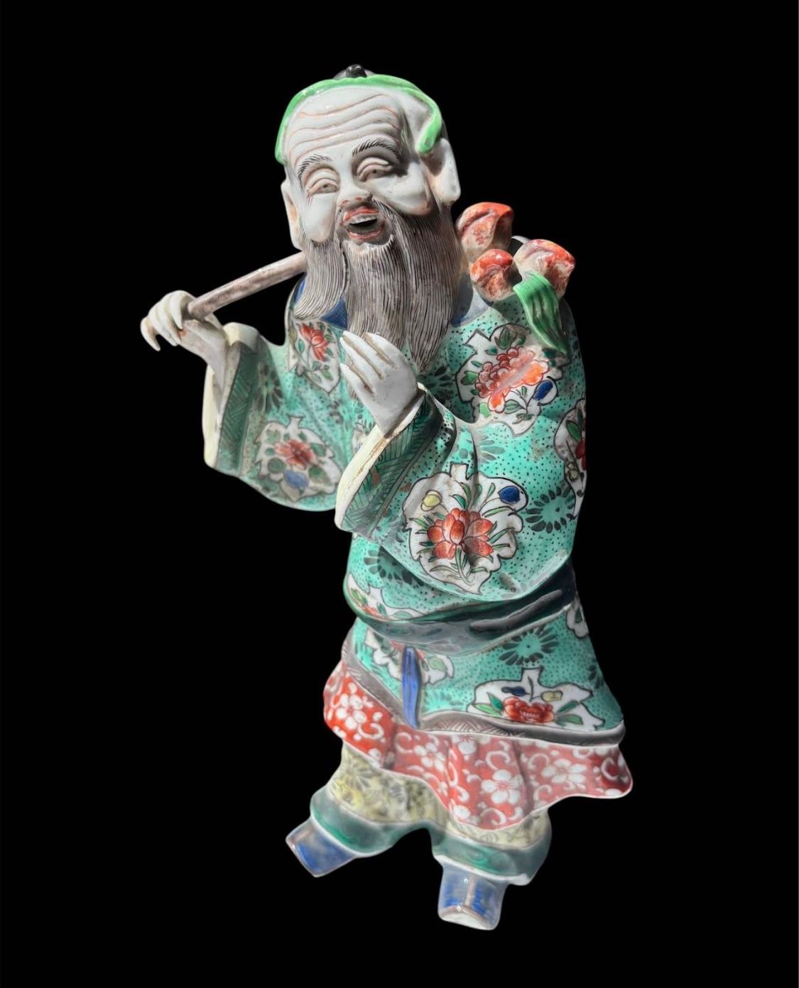 Figurine en céramique représentant un vieil homme en robe à motifs, jouant de la flûte et portant des fleurs.