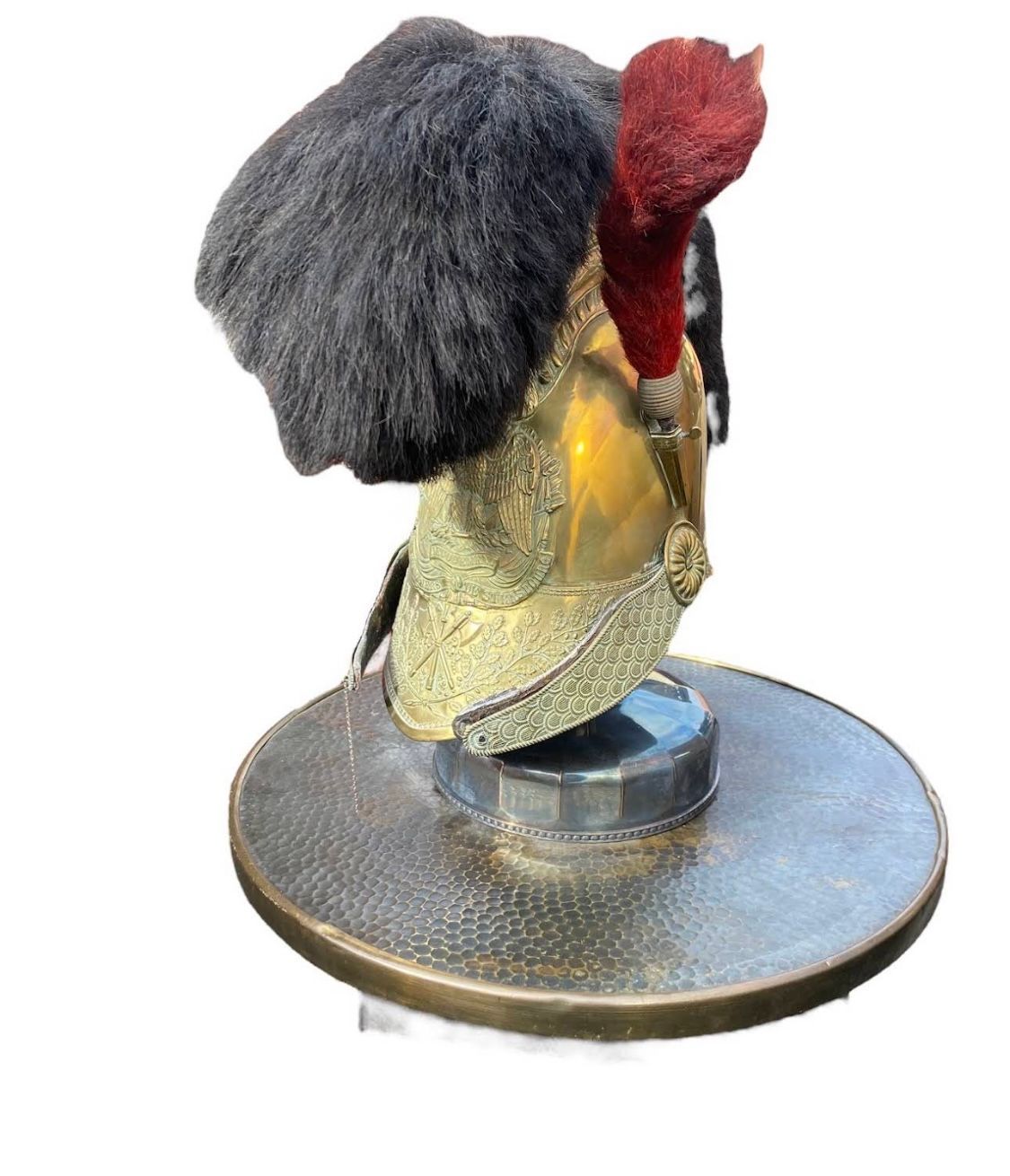 Casque militaire en laiton avec crête en fourrure noire et panache rouge, sur un support rond.