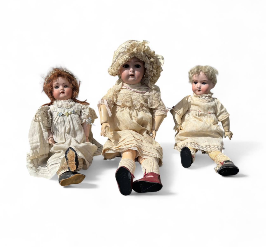 Trois poupées anciennes en porcelaine, vêtues de vêtements d'époque, assises sur du blanc.