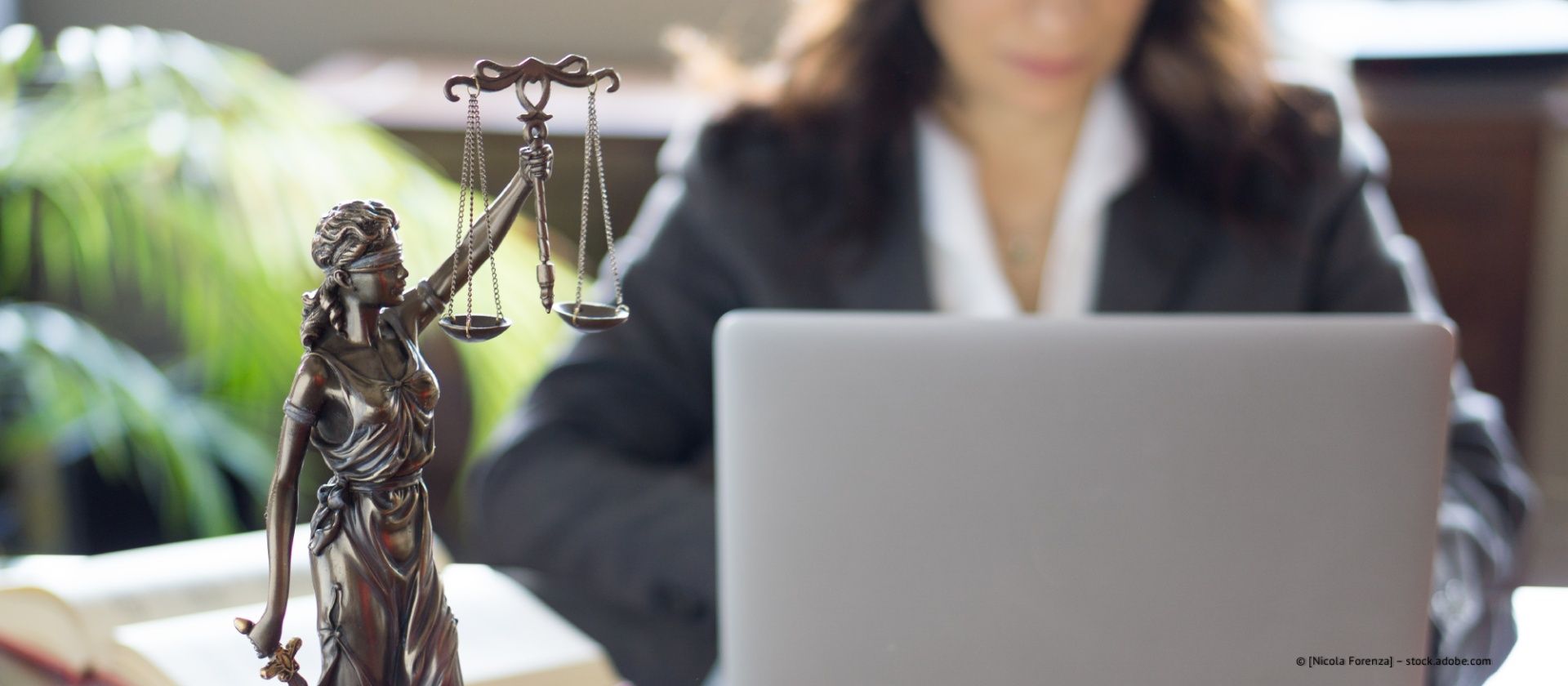 Justitiastatue neben einem Laptop, im Hintergrund ist eine verschwommene Frau bei der Arbeit zu sehen.