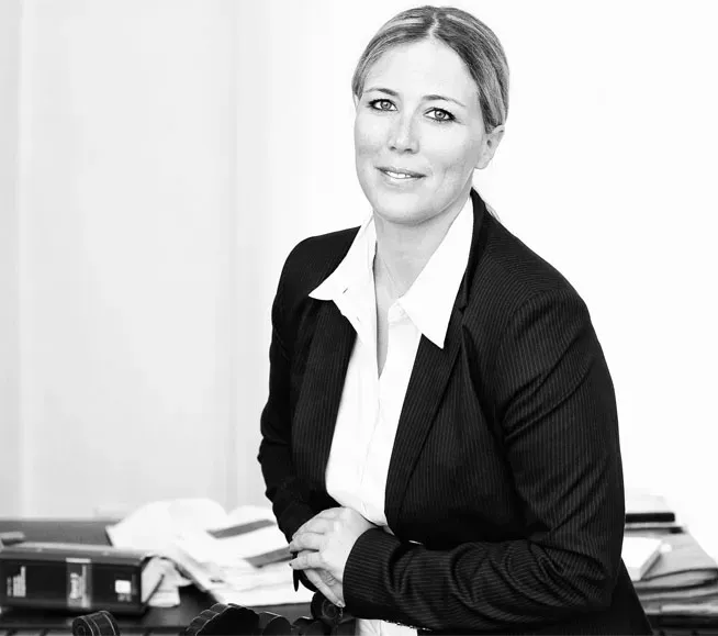 Eine berufstätige Frau in schwarzem Blazer und weißem Hemd sitzt an einem Schreibtisch in einem Büro.