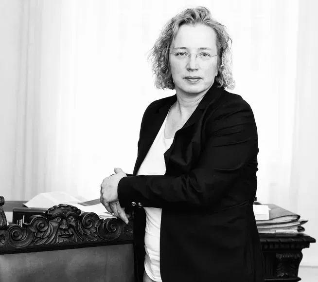 Schwarz-Weiß-Porträt einer Frau, die neben einem reich verzierten Schreibtisch mit Büchern und Papieren steht.