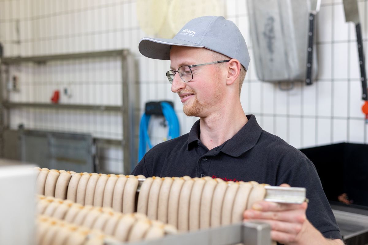 Ein Mann mit Hut und Brille hält in einer Küche frische Bratwurst in der Hand.