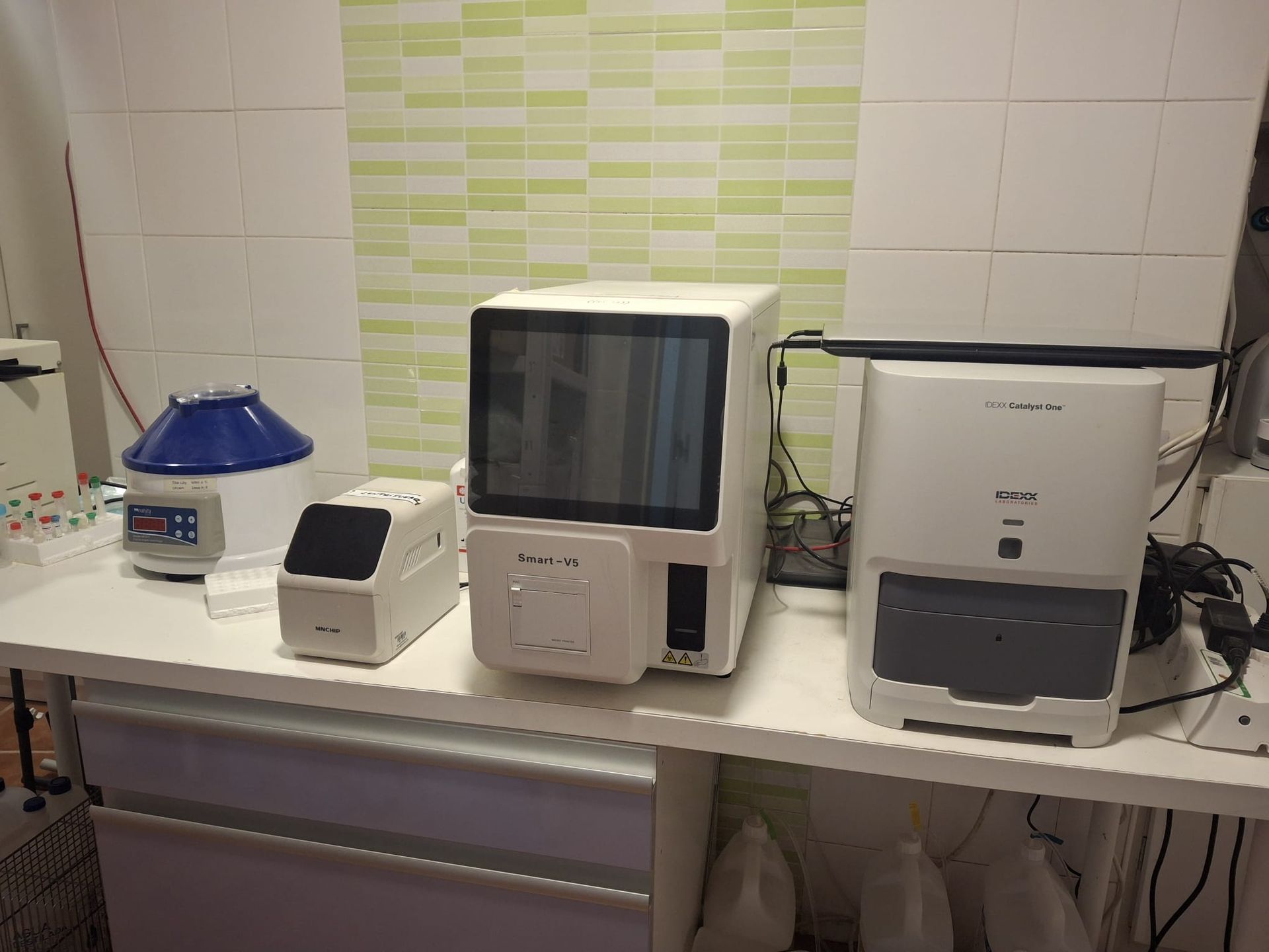 Equipo de laboratorio sobre un mostrador blanco. Incluye una centrífuga, un analizador de sangre y otras máquinas, con una pared de azulejos verdes al fondo.
