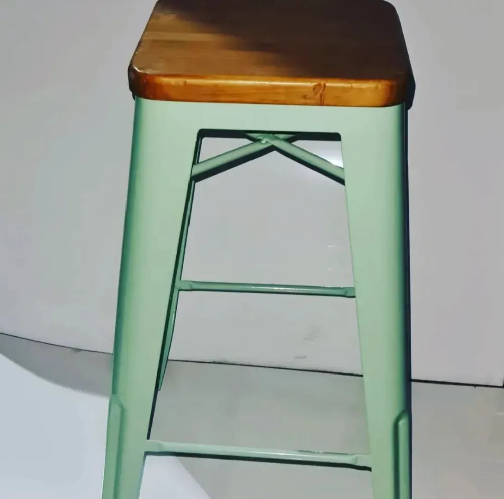 Taburete metálico verde menta con asiento de madera contra una pared blanca lisa.