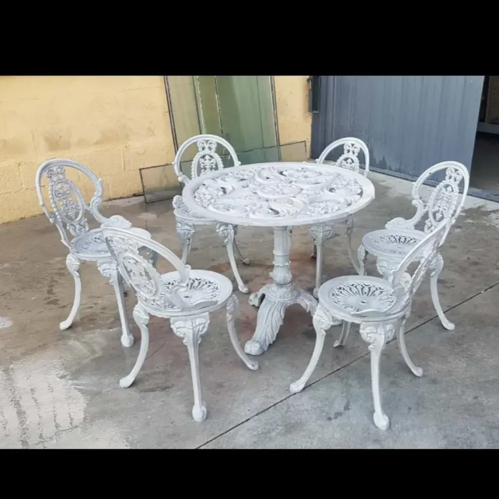 Mesa de patio de metal blanco ornamentado con cinco sillas a juego sobre una superficie de hormigón al aire libre.