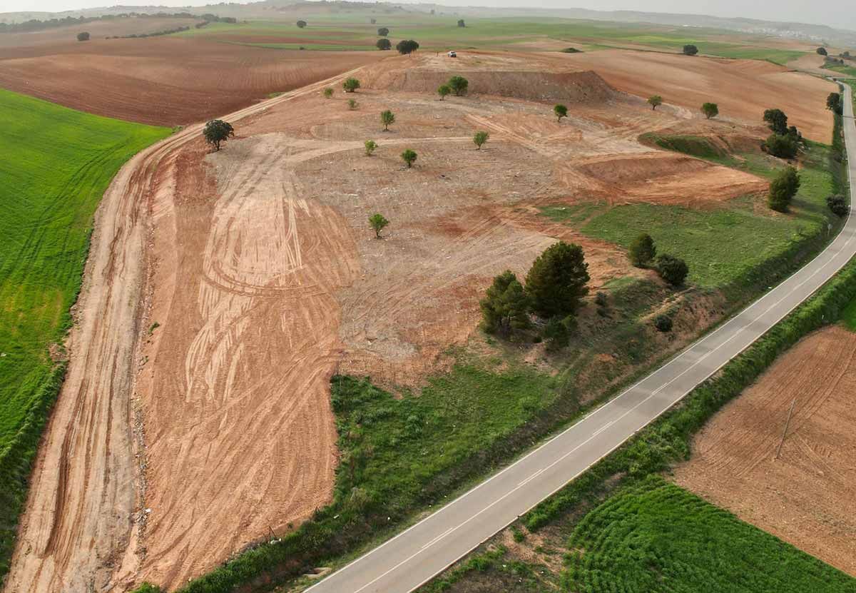Vista aérea de una ladera arada con árboles dispersos junto a un camino rural y campos verdes.