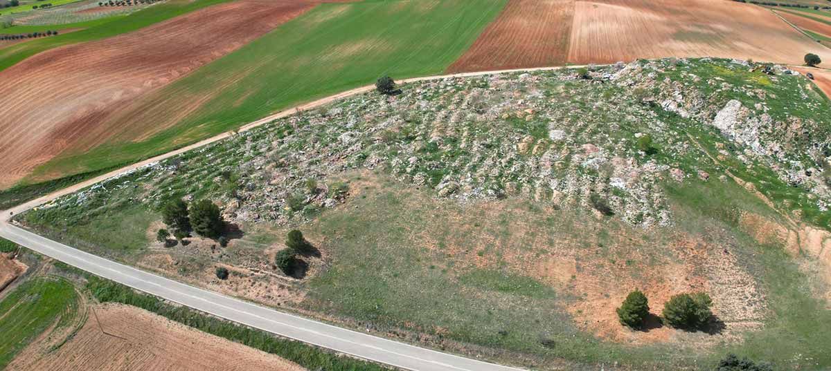 Vista aérea de una ladera cubierta de hierba junto a una carretera y campos de cultivo que forman un mosaico.