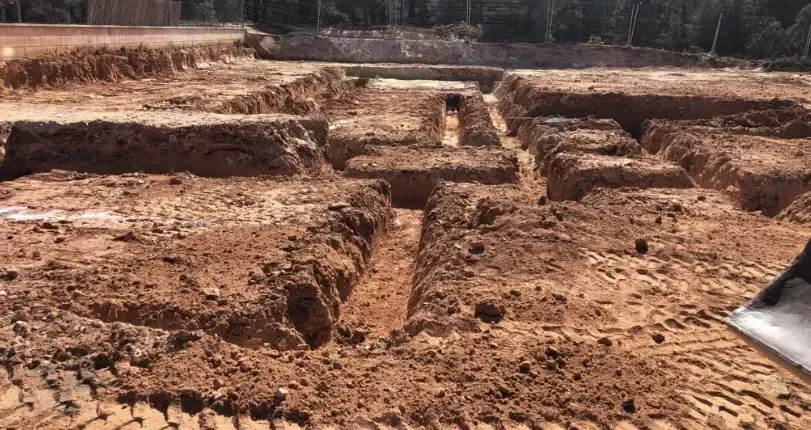 Zanjas excavadas en un terreno despejado con suelo removido y árboles al fondo.