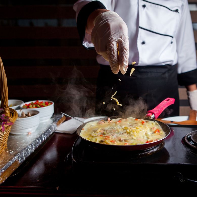 O chef polvilha queijo em uma omelete quente em uma panela, com os ingredientes e a estação de preparação ao lado.