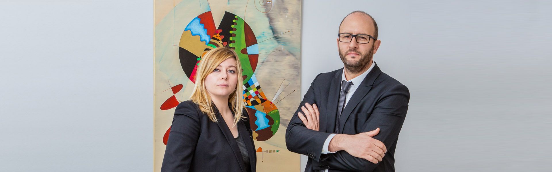 Deux avocats se tiennent à côté d'une sculpture murale abstraite et colorée, l'un d'eux ayant les bras croisés.