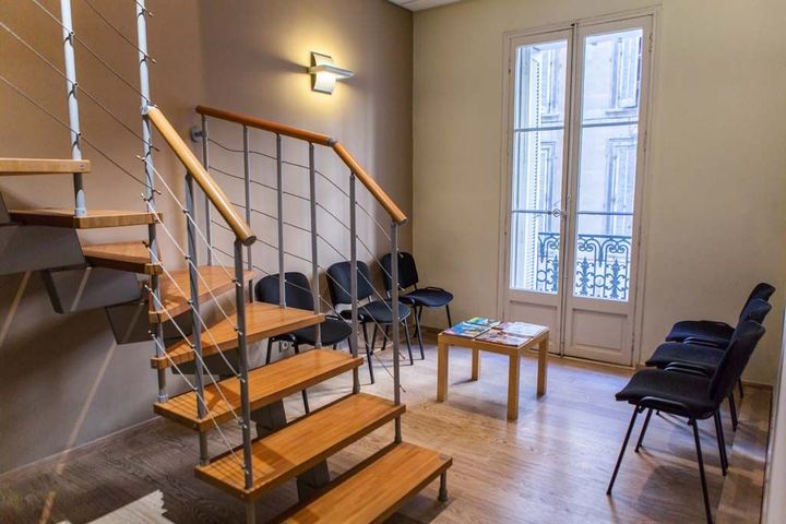 Salle d'attente vide avec escalier, chaises noires, petite table et hautes fenêtres à rideaux bleus.
