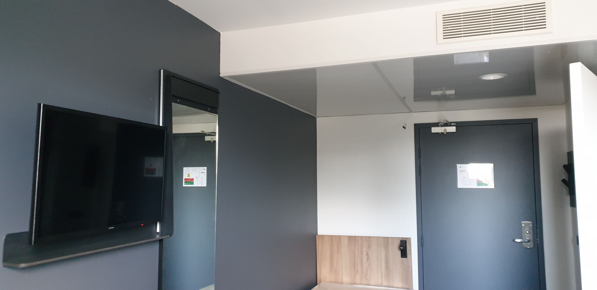 Une chambre d'hôtel avec une télévision fixée au mur, un miroir et une porte fermée. Murs gris et plafond clair.