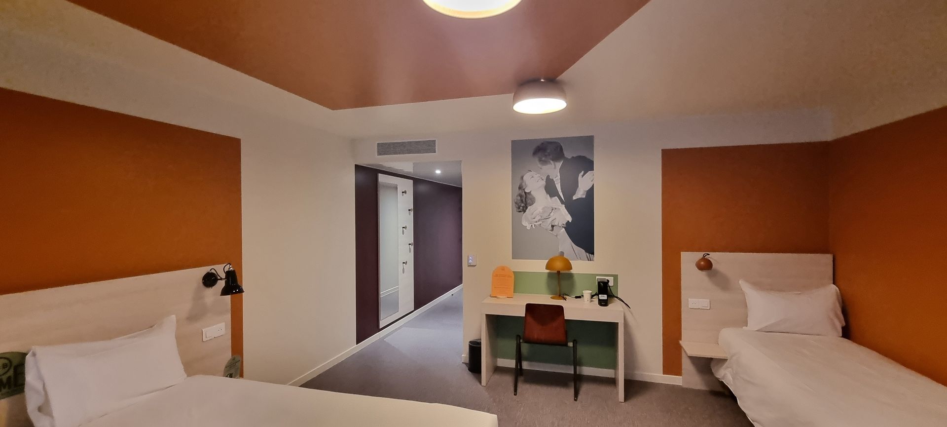 Chambre d'hôtel avec deux lits, un bureau et un couloir. Murs orange et blancs, moquette grise.