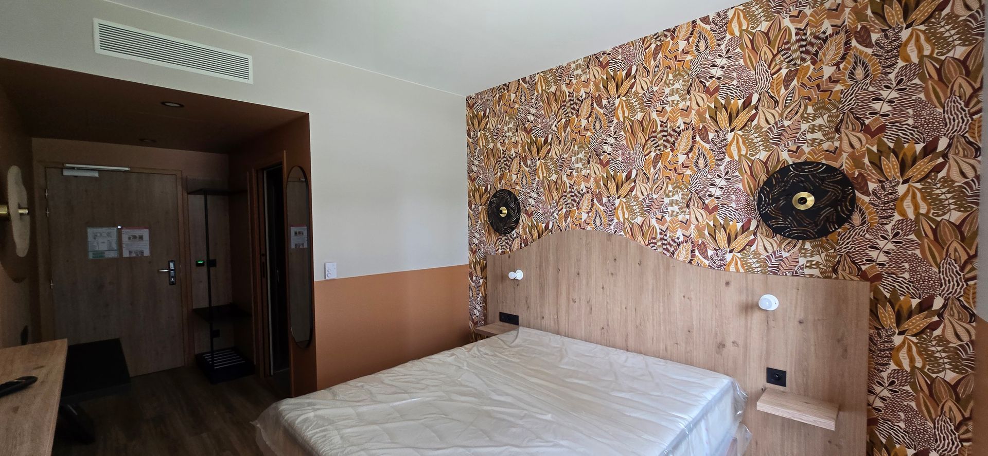 Chambre avec lit adossé à un mur à motifs de feuilles. Tête de lit en bois clair et petites étagères. Porte à gauche.