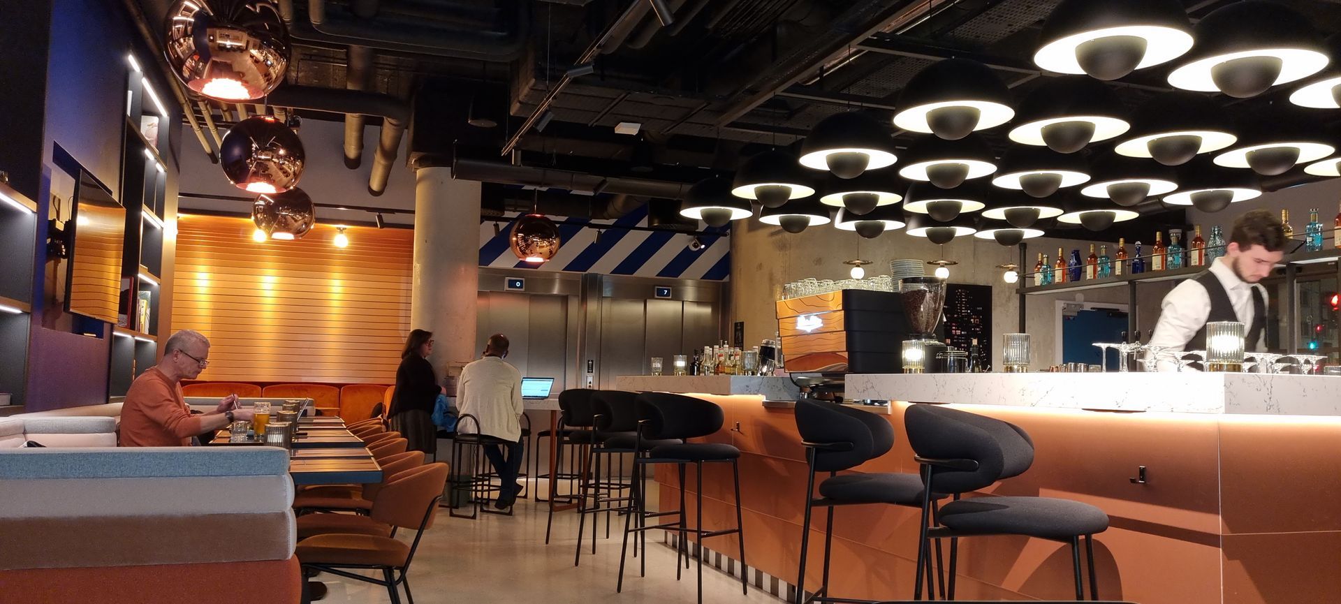 Intérieur de bar avec des clients assis, un barman à l'œuvre et un décor en cuivre et blanc.