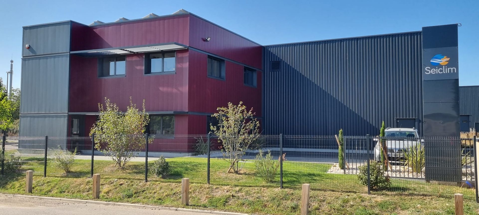 Bâtiment aux panneaux bordeaux et gris foncé, enseigne noire avec logo, voiture blanche derrière un portail.