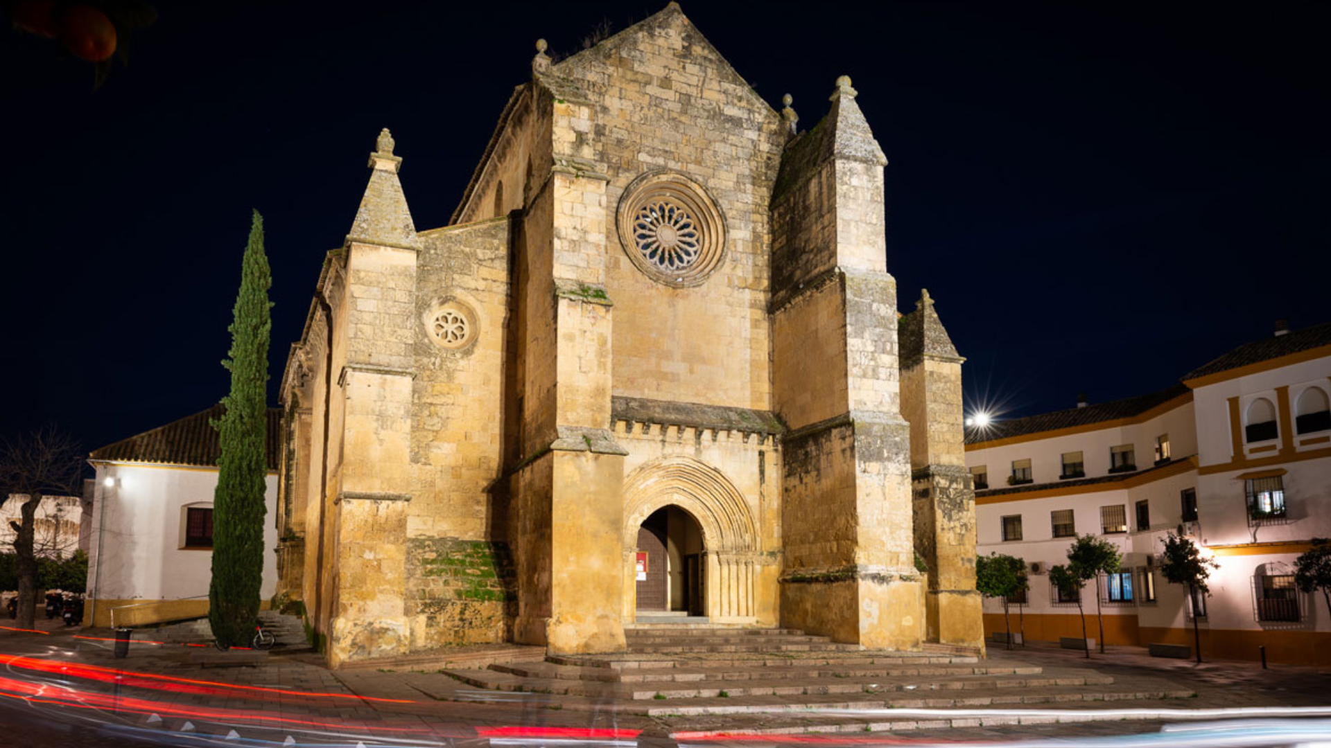 Fachada de iglesia de piedra iluminada por la noche, con un rosetón y escalones que conducen a la entrada.