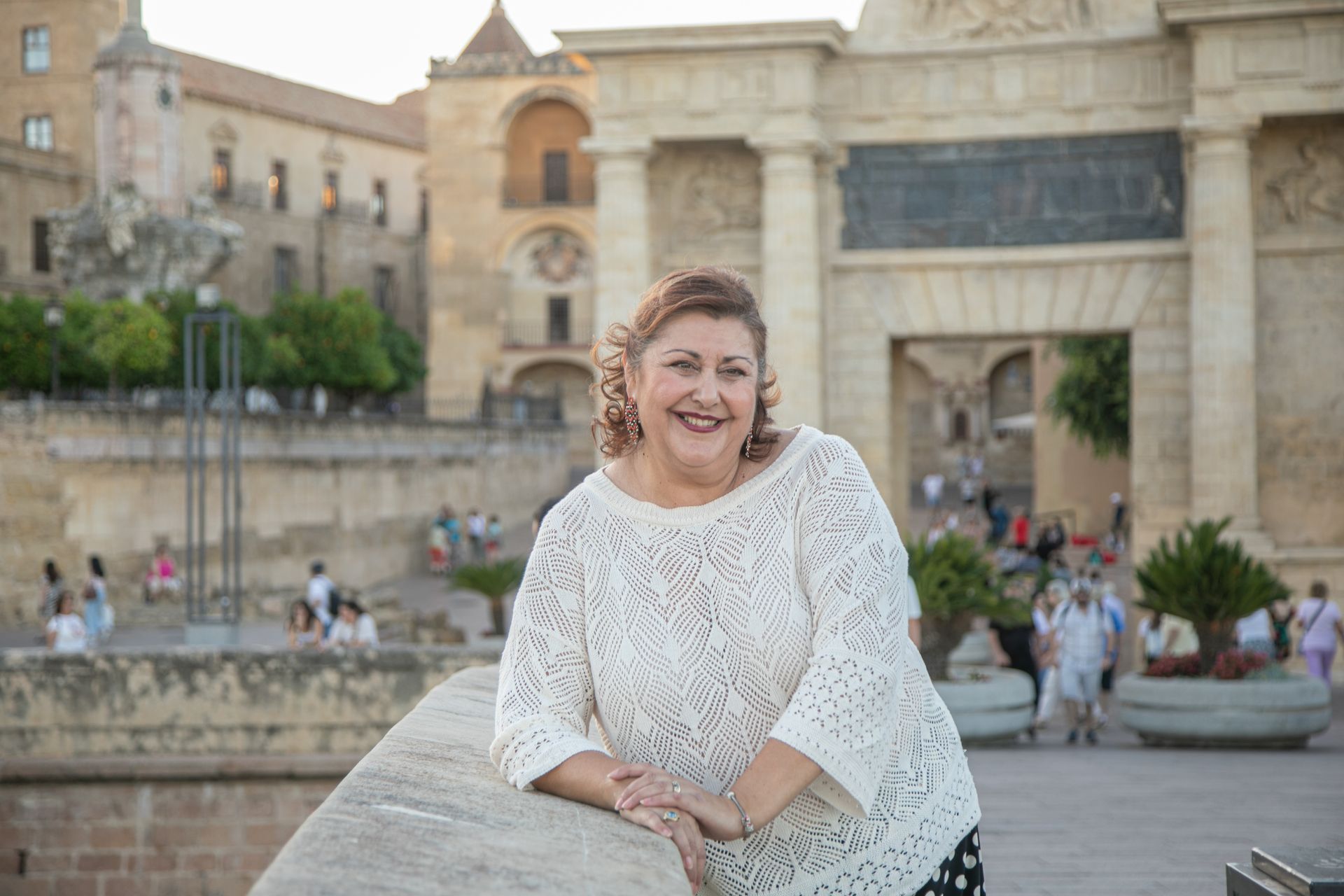Mujer con suéter blanco sonríe junto a un puente de piedra y un arco en Córdoba, España.