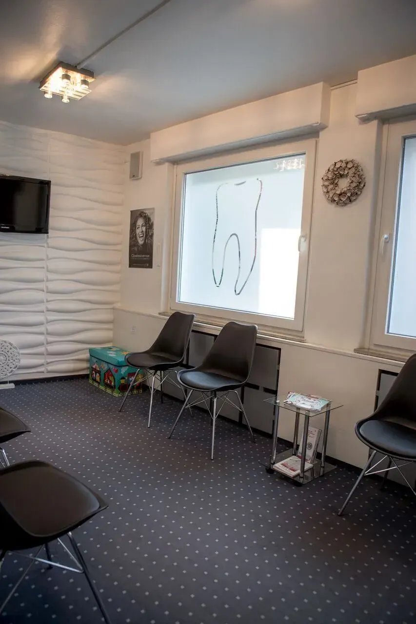 Leeres Wartezimmer mit grauen Stühlen, gemustertem Teppichboden, großen Fenstern und einer Wandleuchte.