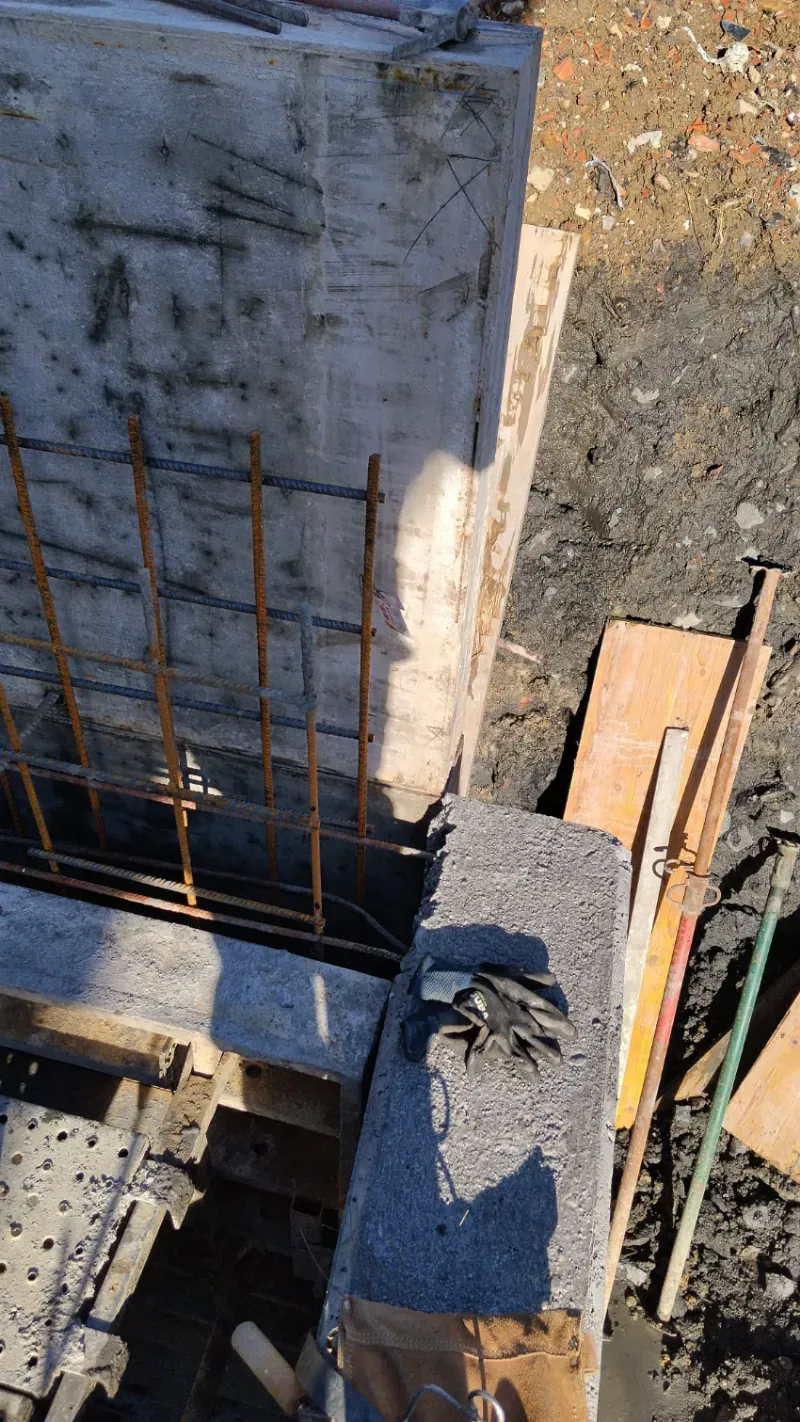 Obra en construcción con muro de hormigón, barras de refuerzo y tablones de madera alrededor de una columna vertical.