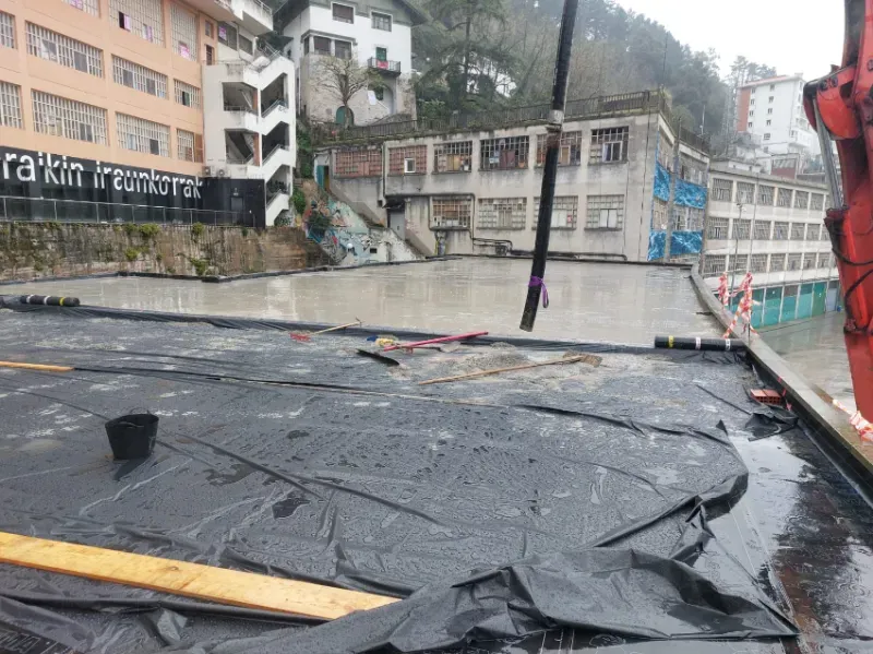 Calle inundada con escombros de lona negra derrumbada junto a un edificio dañado en un pueblo de montaña lluvioso.