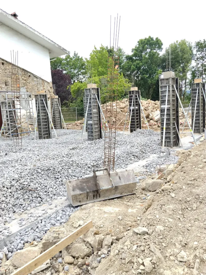 Obra en construcción con pilares de cimentación de hormigón y barras de refuerzo sobre grava junto a un edificio.