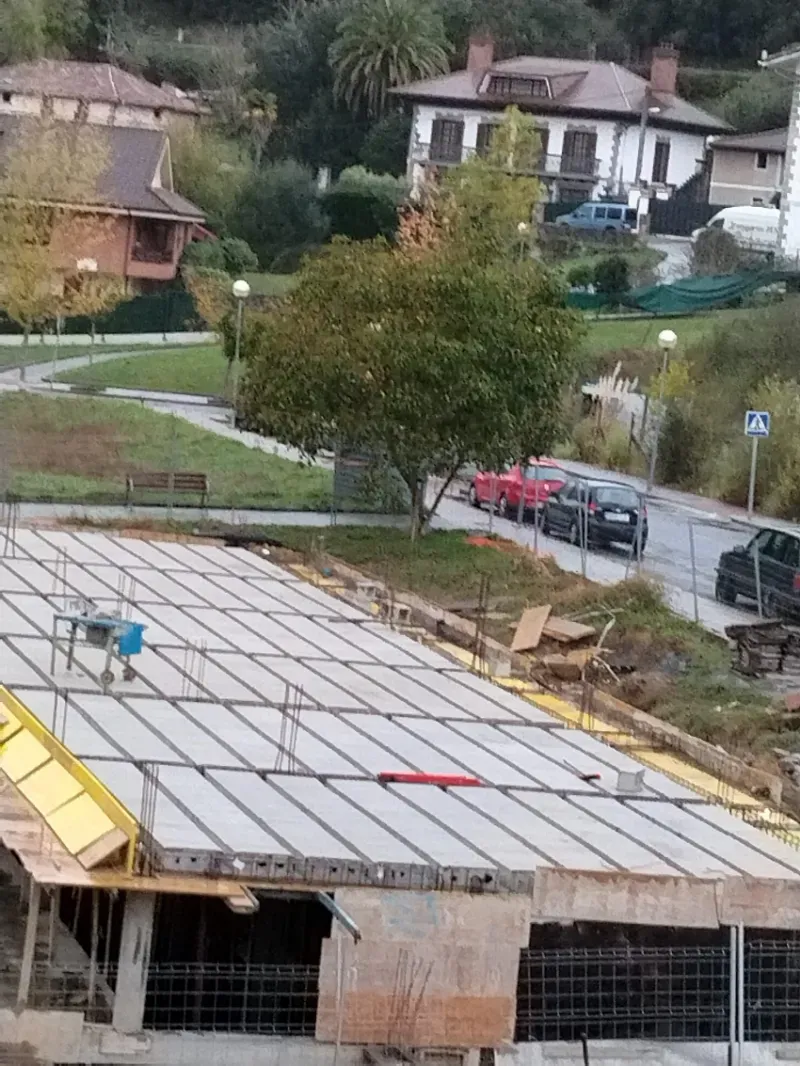 Obra en construcción con cimientos de una casa, rejilla de varillas de refuerzo y una carretera en pendiente con vehículos estacionados en las cercanías.