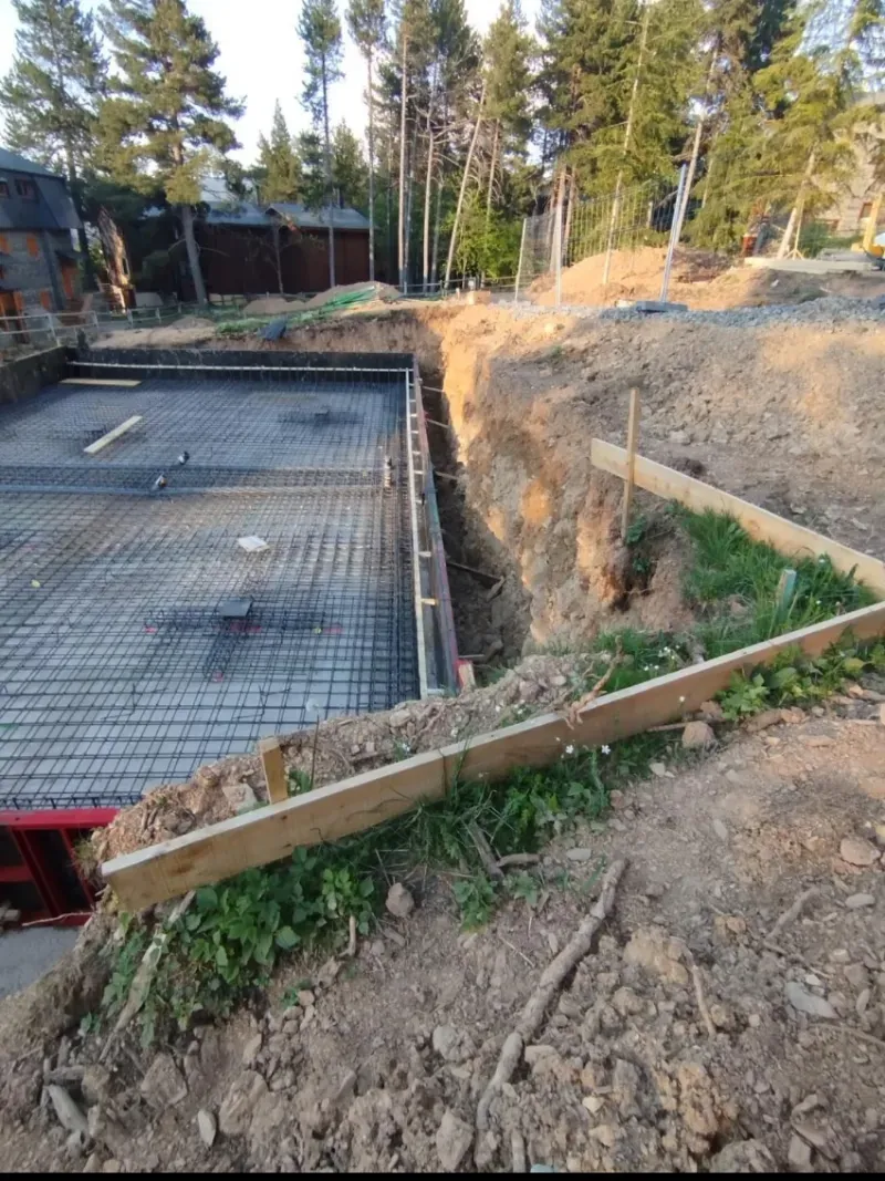 Obra en construcción con cimientos de hormigón, barras de refuerzo, terraplén de tierra y árboles al fondo.