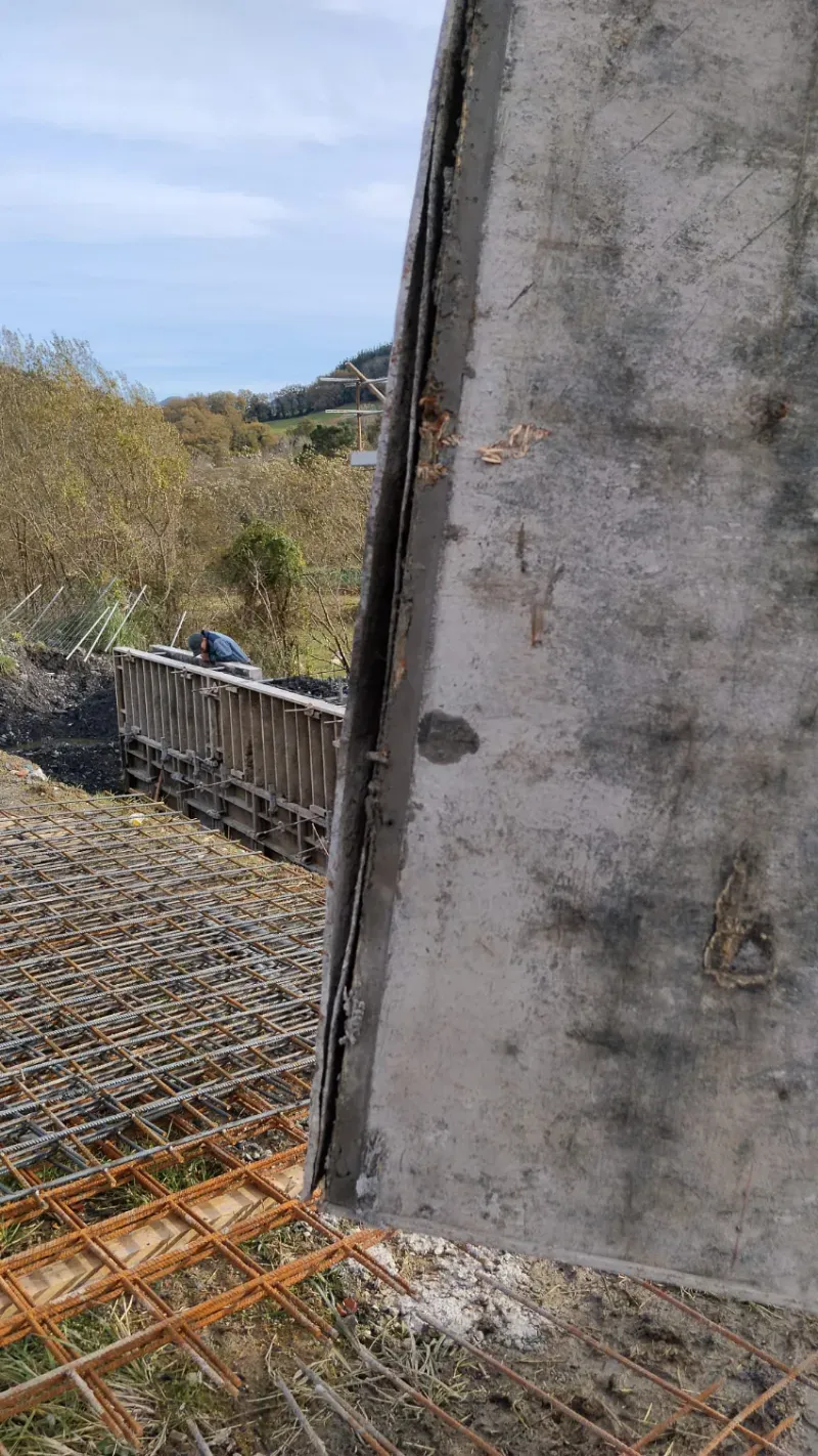 Muro de hormigón junto a una cimentación de barras de refuerzo en una obra en construcción cerca de hierba alta y cielo abierto.
