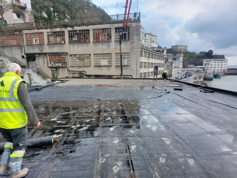 Trabajadores reparando un paseo marítimo dañado junto a edificios y agua.